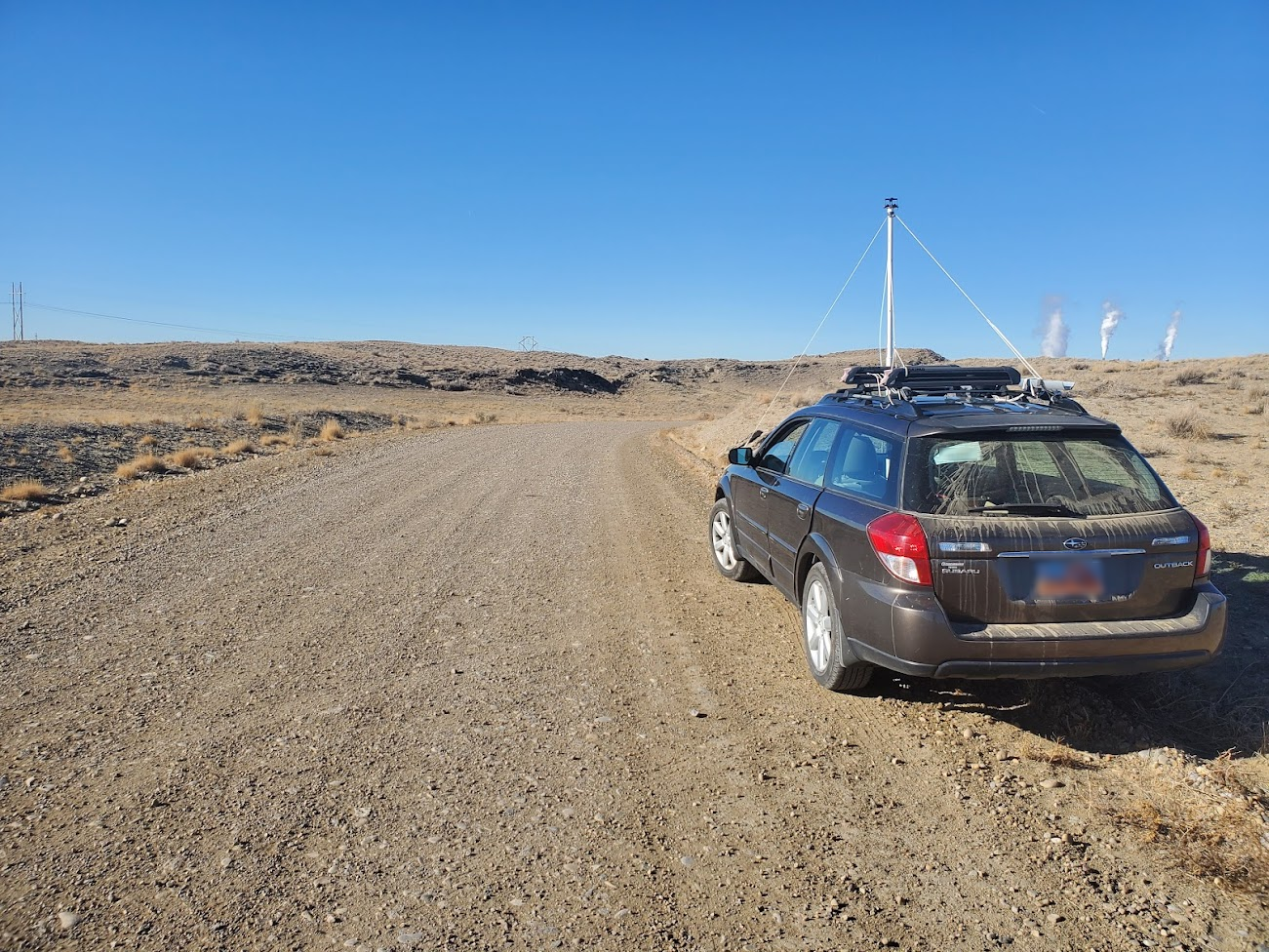 A hatchback car is parked on the side of a dirt road adjacent to short grasses. The roof of the car is equipped with environmental monitoring equipment, including a TriSonica<sup>&reg;</sup> Mini Wind and Weather Sensor and an air intake for a trace gas analyzer. An industrial facility or oil refinery is visible on the distant horizon.